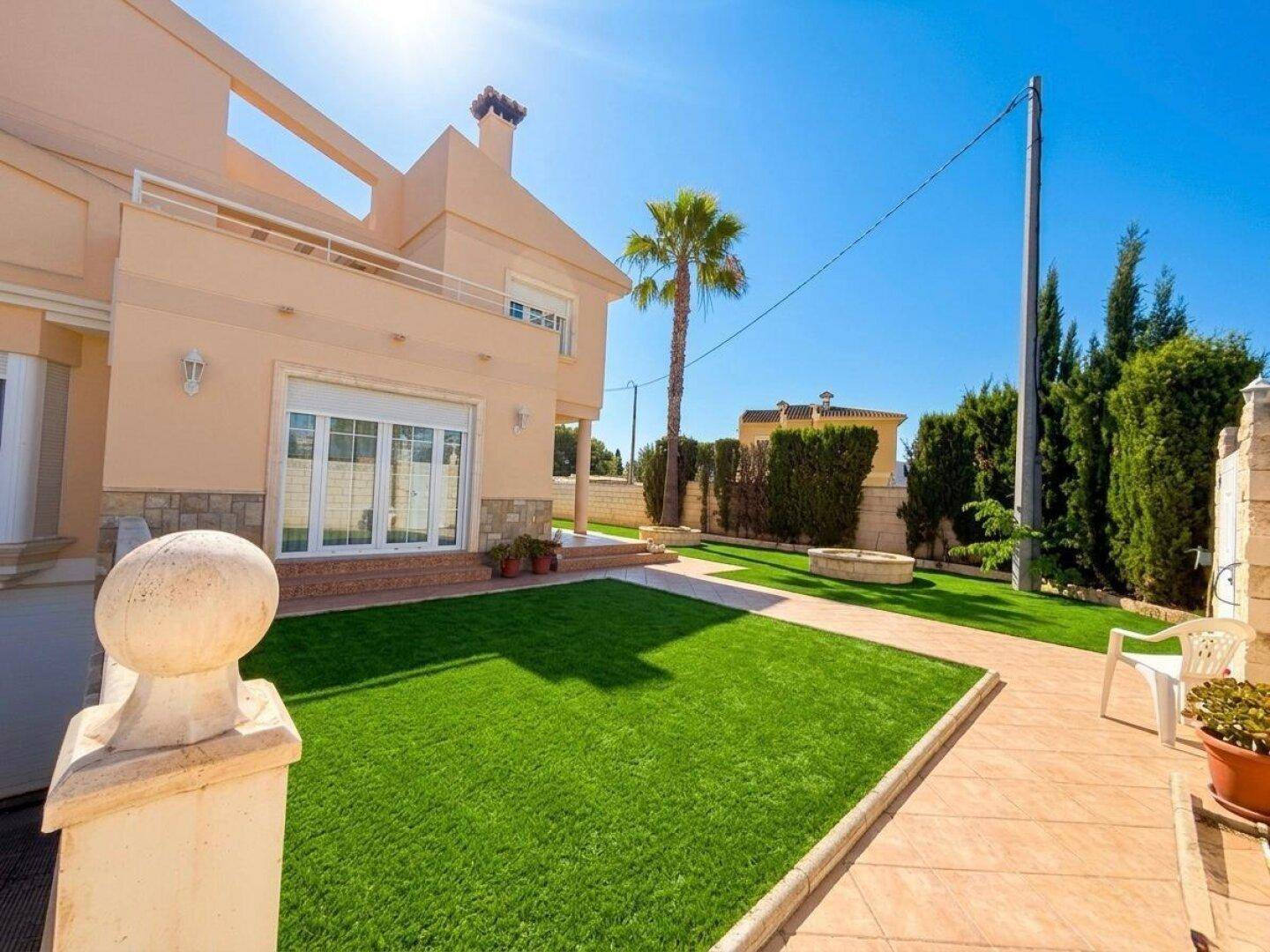 Rückansicht des Hauses mit grünem Rasen, gefliester Terrasse und mediterraner Bepflanzung.