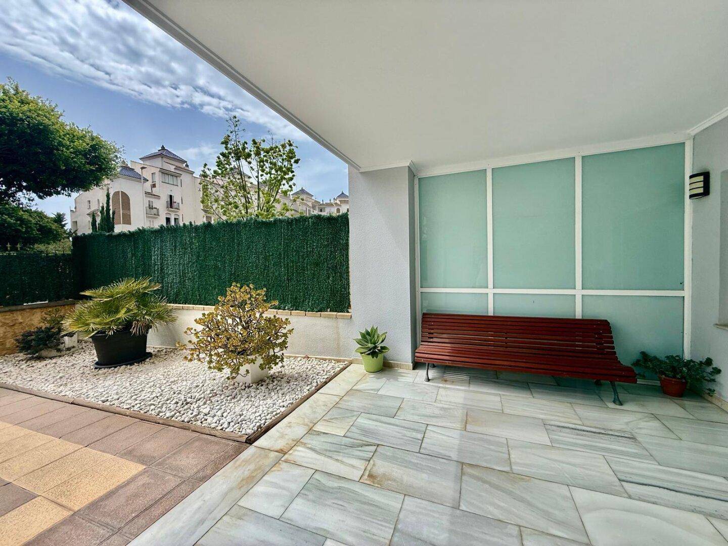Überdachte Terrasse mit Marmorfliesen, Sitzbank und einem kleinen, pflegeleichten Gartenbereich.