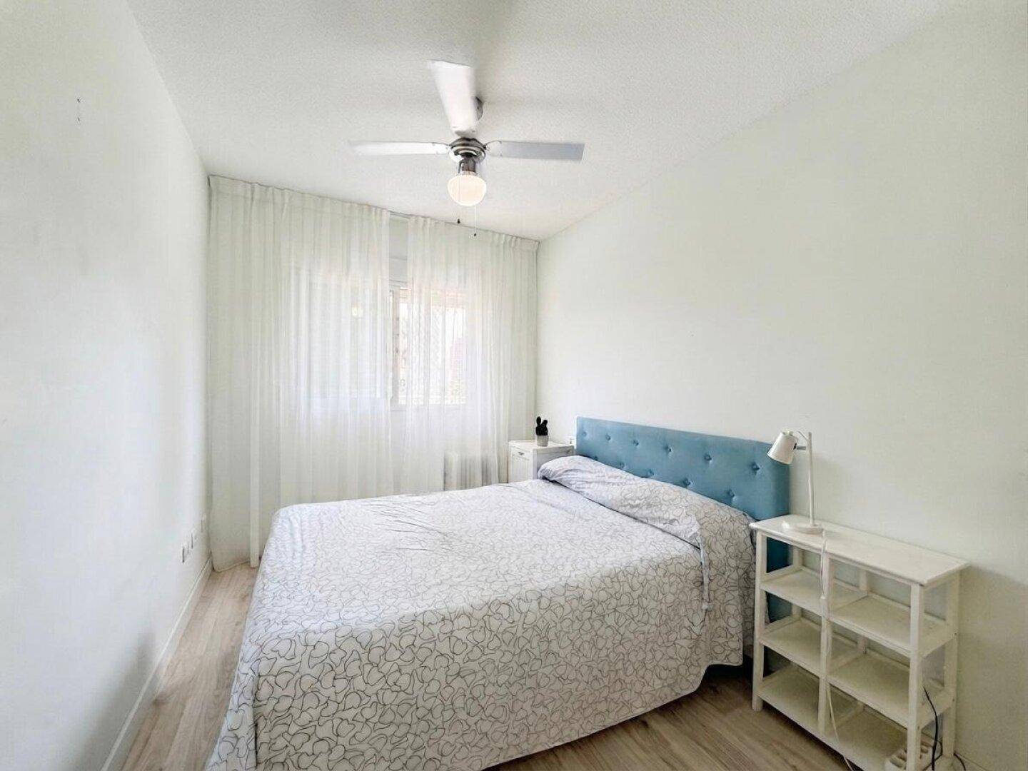 Helles Schlafzimmer mit Doppelbett, blauem Kopfteil, weißen Wänden und Deckenventilator.