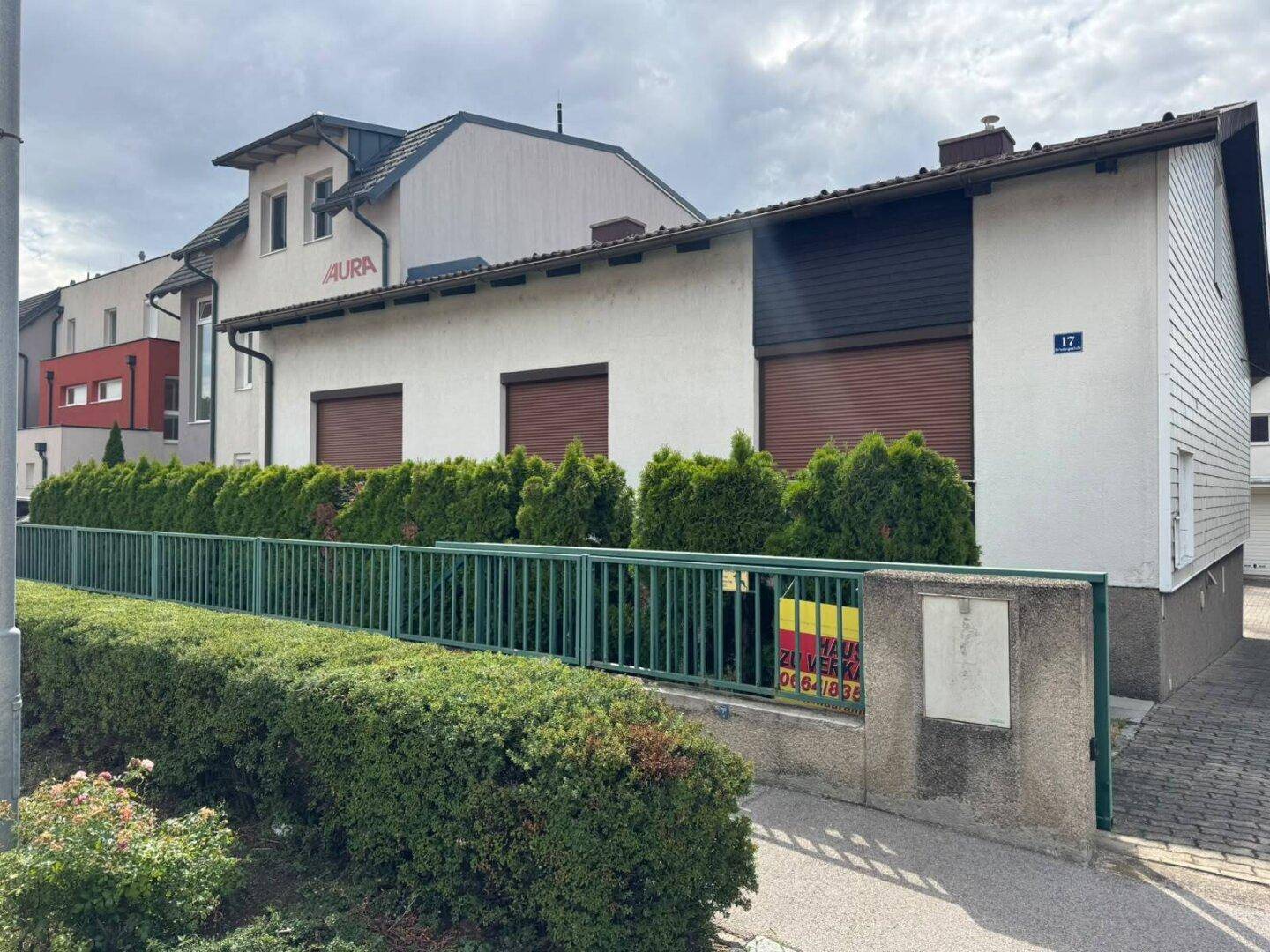 Ansicht des Hauses von der Straße mit grüner Hecke und Rollläden an den Fenstern.