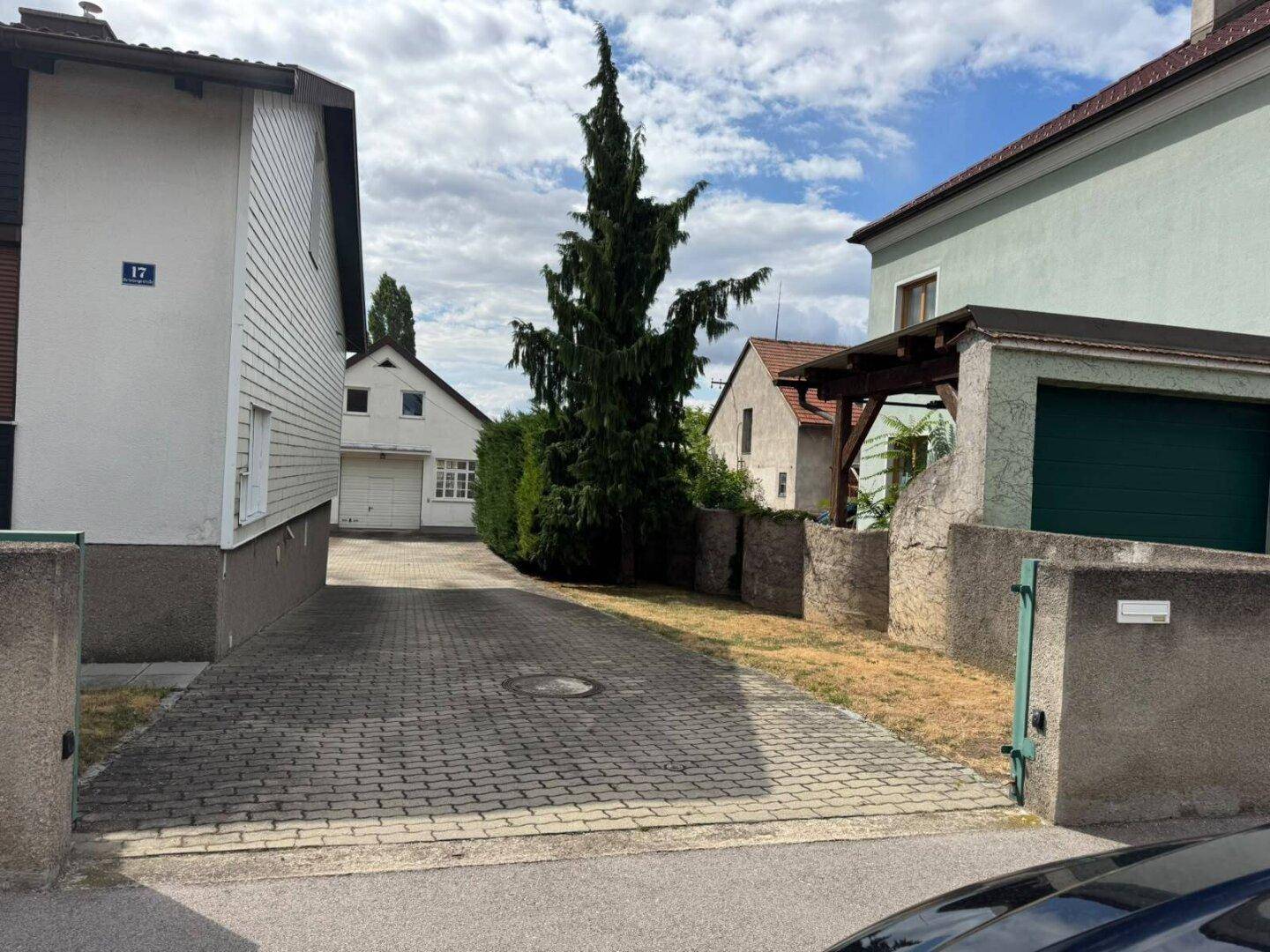 Gepflasterte Einfahrt zwischen den Gebäuden mit hohen Bäumen im Hintergrund.