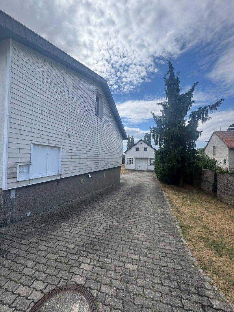 Seitenansicht des Hauses mit gepflasterter Einfahrt und einem weiteren Gebäude im Hintergrund.