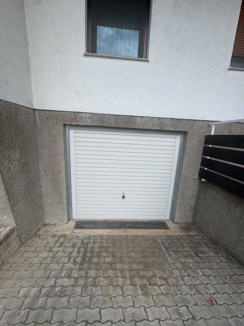 Weiße Garagentür im Untergeschoss des Hauses, zugänglich über die gepflasterte Einfahrt.