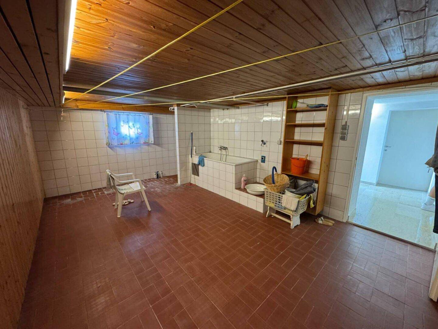 Großer Kellerraum mit Fliesenboden, Holzdecke und einer Badewanne im hinteren Bereich.