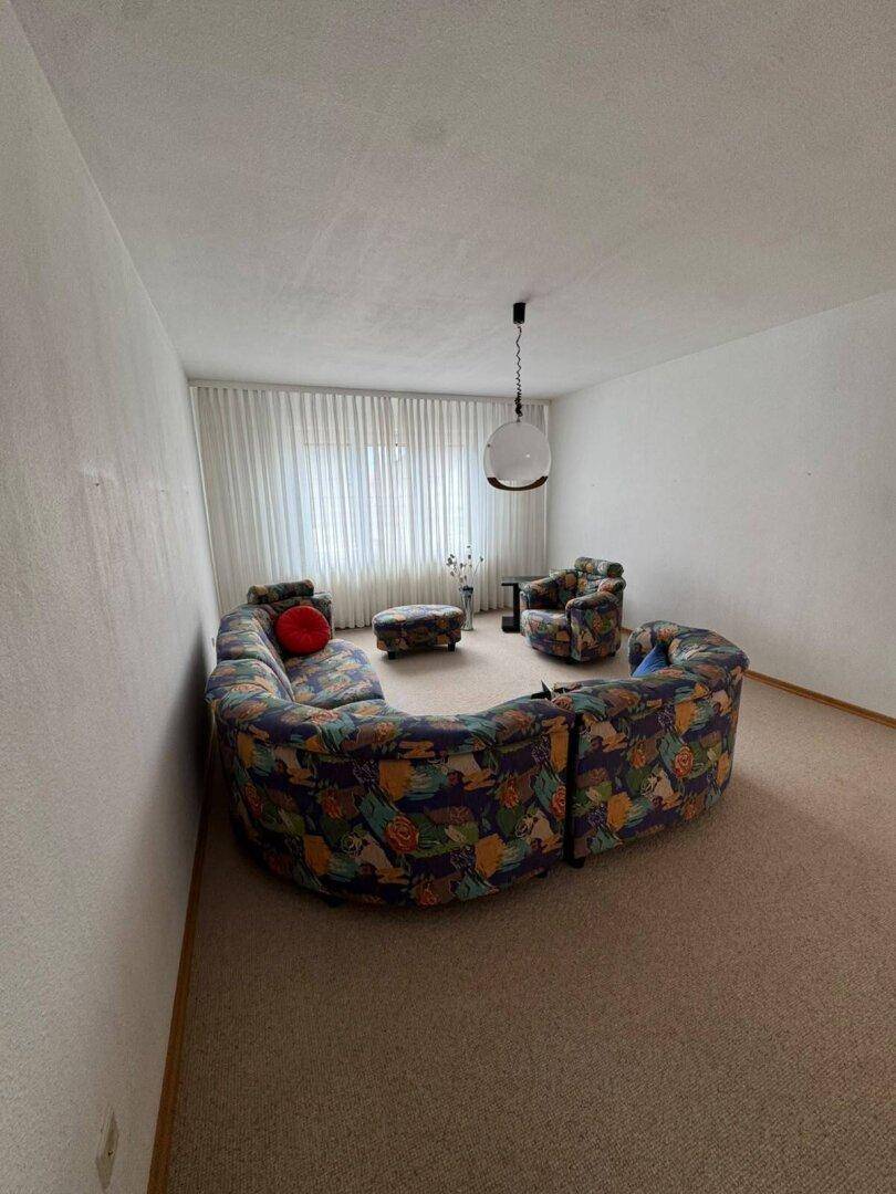 Helles Wohnzimmer mit großem, gemustertem Sofa und Fensterfront, bietet viel Tageslicht.