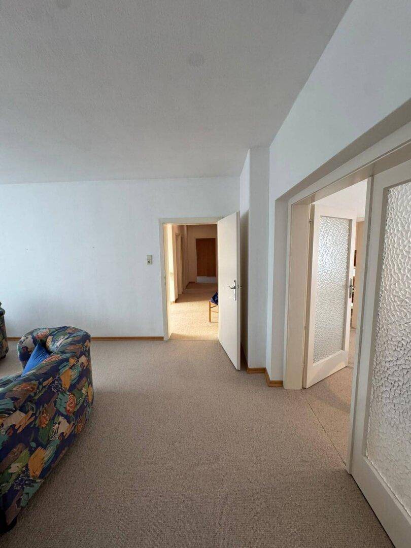 Geräumiges Wohnzimmer mit Teppichboden und Zugang zu weiteren Räumen, hell und funktional.