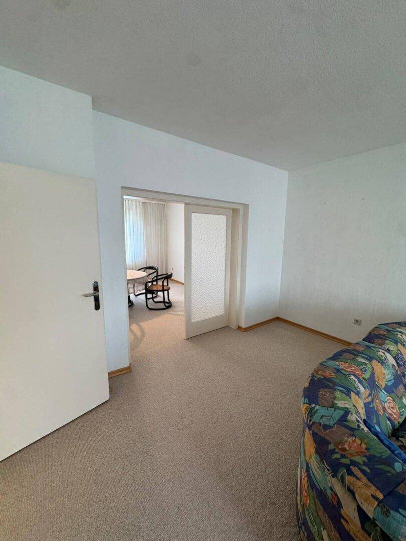 Wohnzimmer mit Teppichboden und Durchgang zu einem angrenzenden Essbereich, schlicht eingerichtet.
