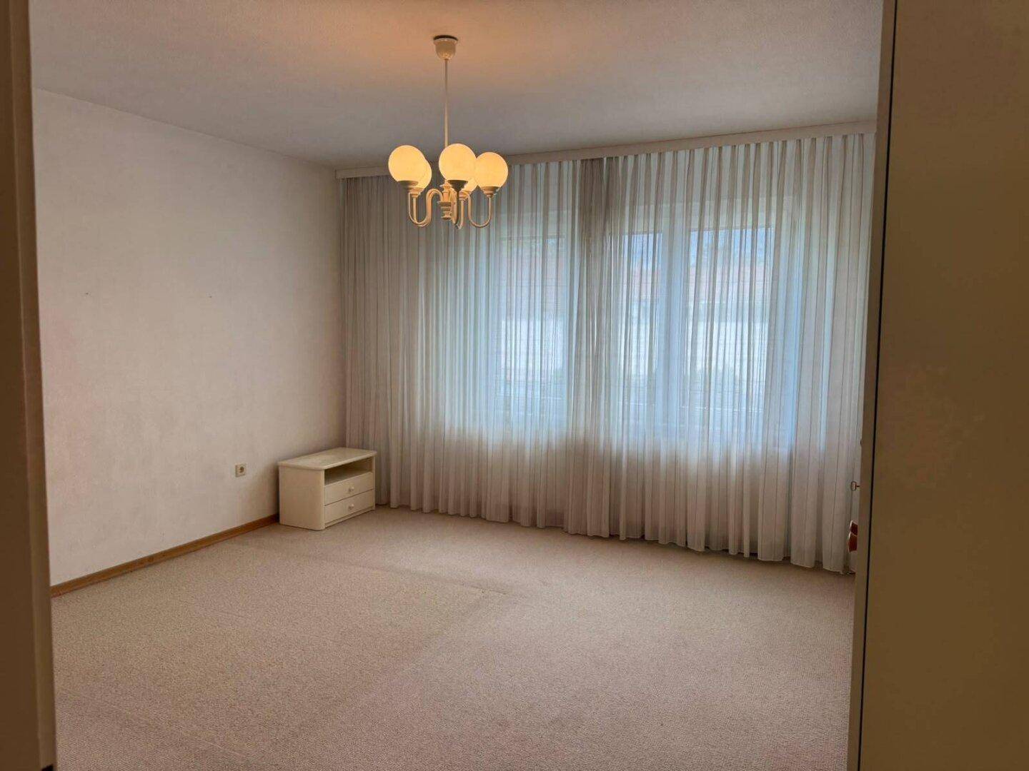 Helles Schlafzimmer mit Teppichboden, großem Fenster und klassischem Kronleuchter, einfach eingerichtet.