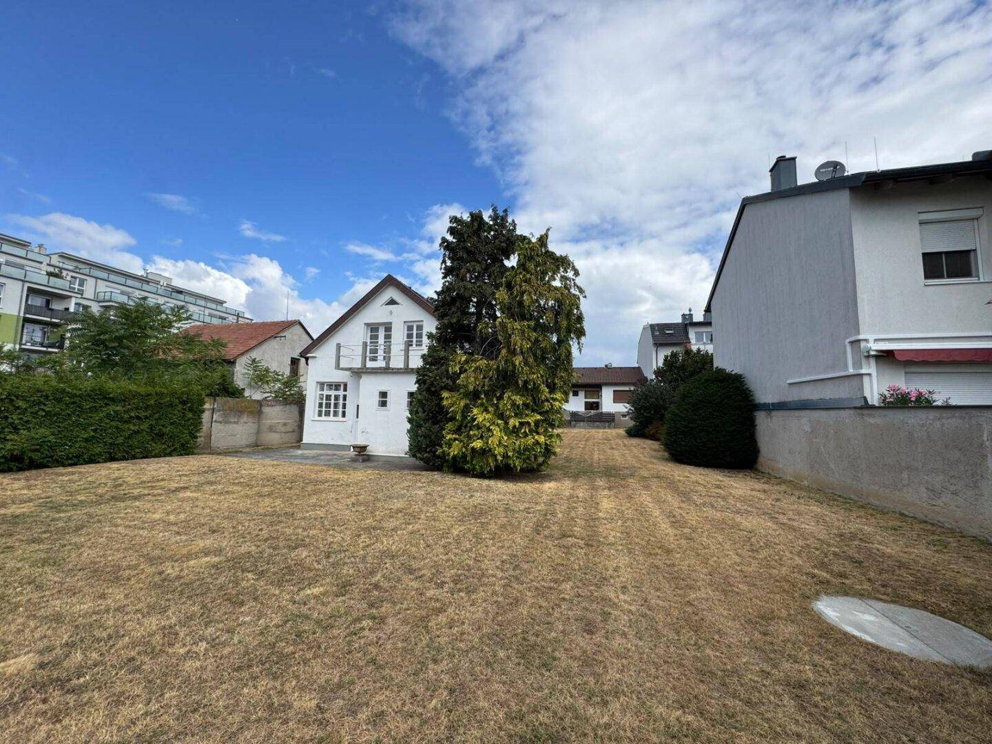 Rückansicht des Hauses mit trockenem Rasen und hohen Bäumen, unter blauem Himmel.
