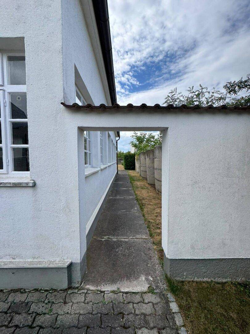 Schmaler Durchgang zwischen Gebäudeteilen mit Betonweg, führt in den hinteren Bereich.