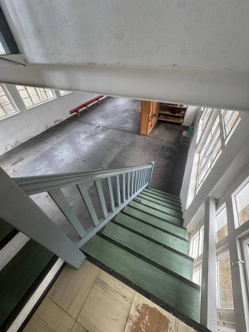 Blick von der Treppe in einen großen, leeren Raum mit Betonboden und vielen Fenstern.