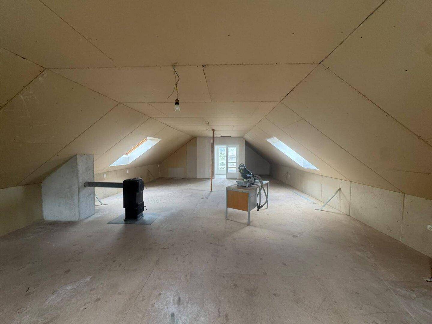 Geräumiger Dachboden im Rohbau mit Dachfenstern, Holzofen und Zugang zu einem weiteren Raum.