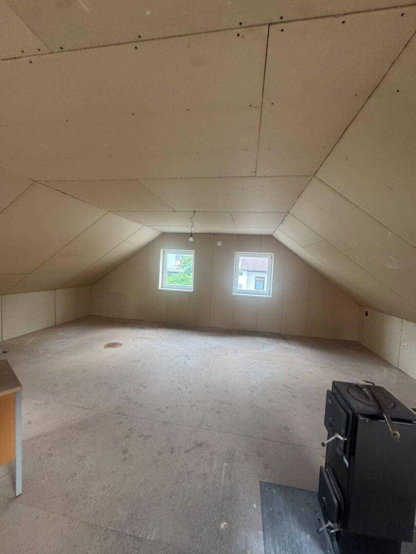 Dachboden im Rohbau mit schrägen Wänden, zwei Fenstern und einem Holzofen.