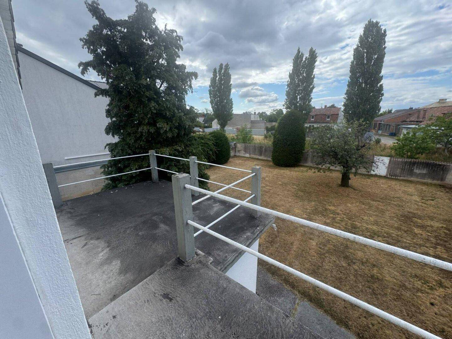 Blick von einem Balkon auf einen trockenen Garten mit Bäumen und umliegenden Häusern.