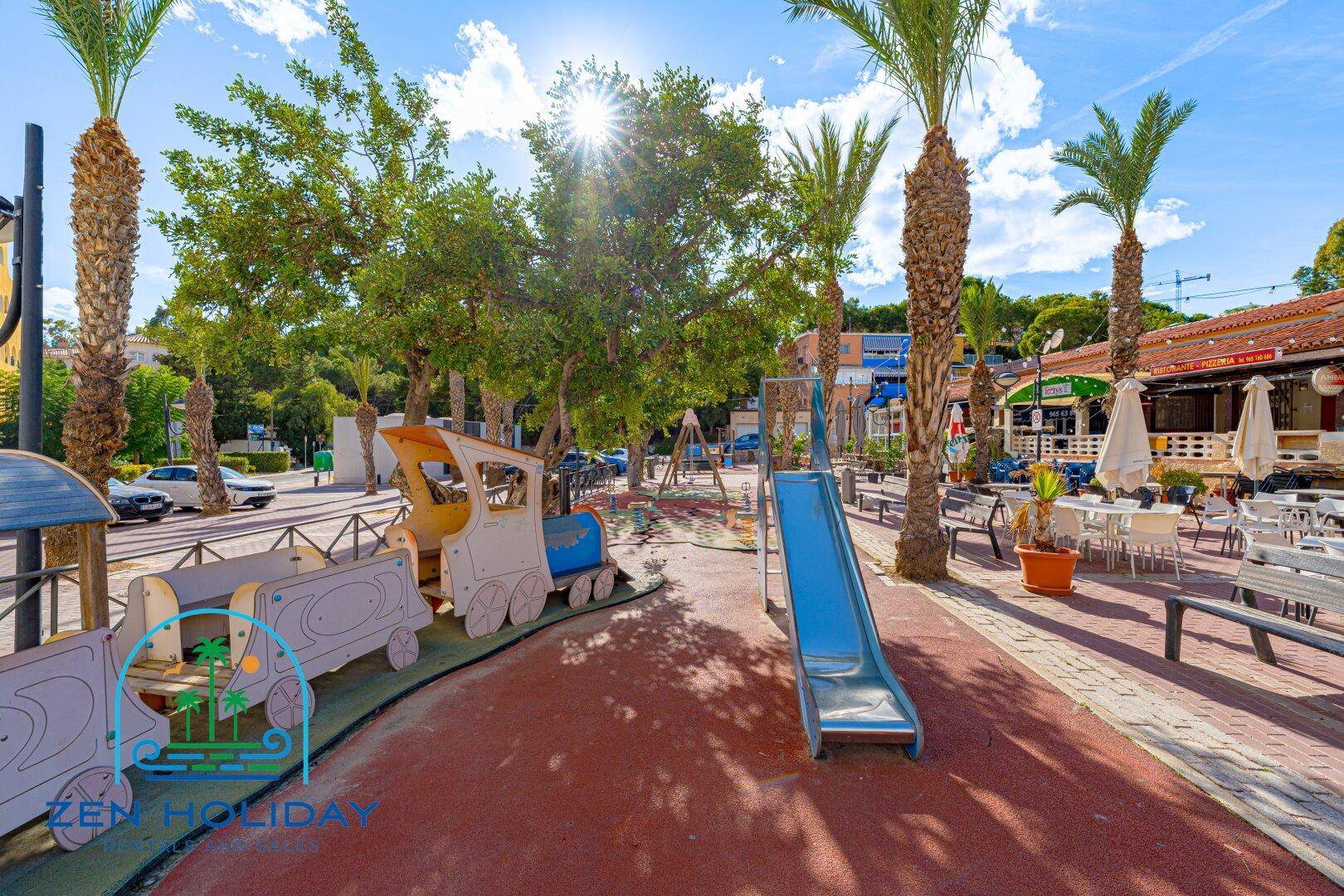 Bunter Spielplatz mit Rutsche und Zug-Spielgerät, umgeben von Palmen und Grünflächen.