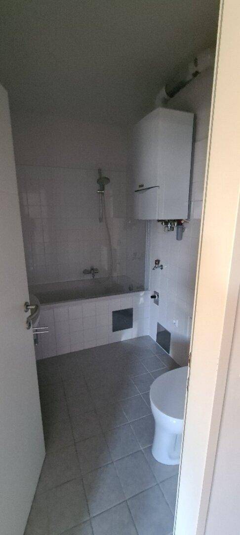 Badezimmer mit Badewanne, Toilette und hellen Fliesen, inklusive Warmwasserbereiter.