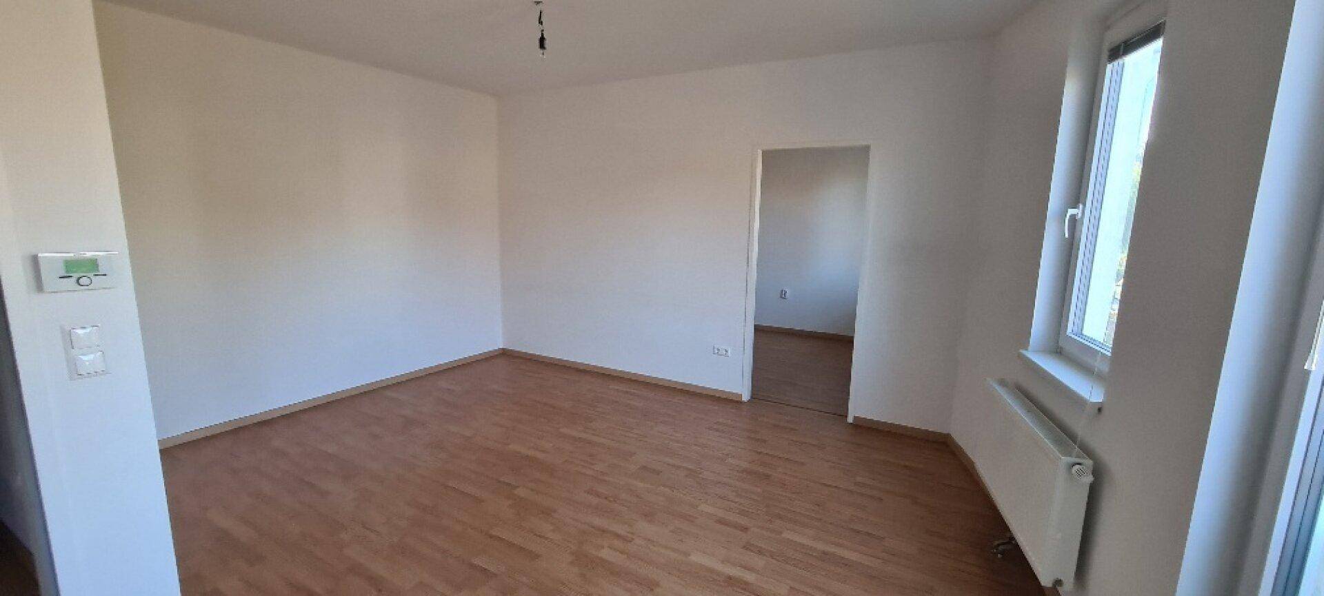 Helles, leeres Zimmer mit Holzboden, weißen Wänden und einem Fenster mit Heizkörper.