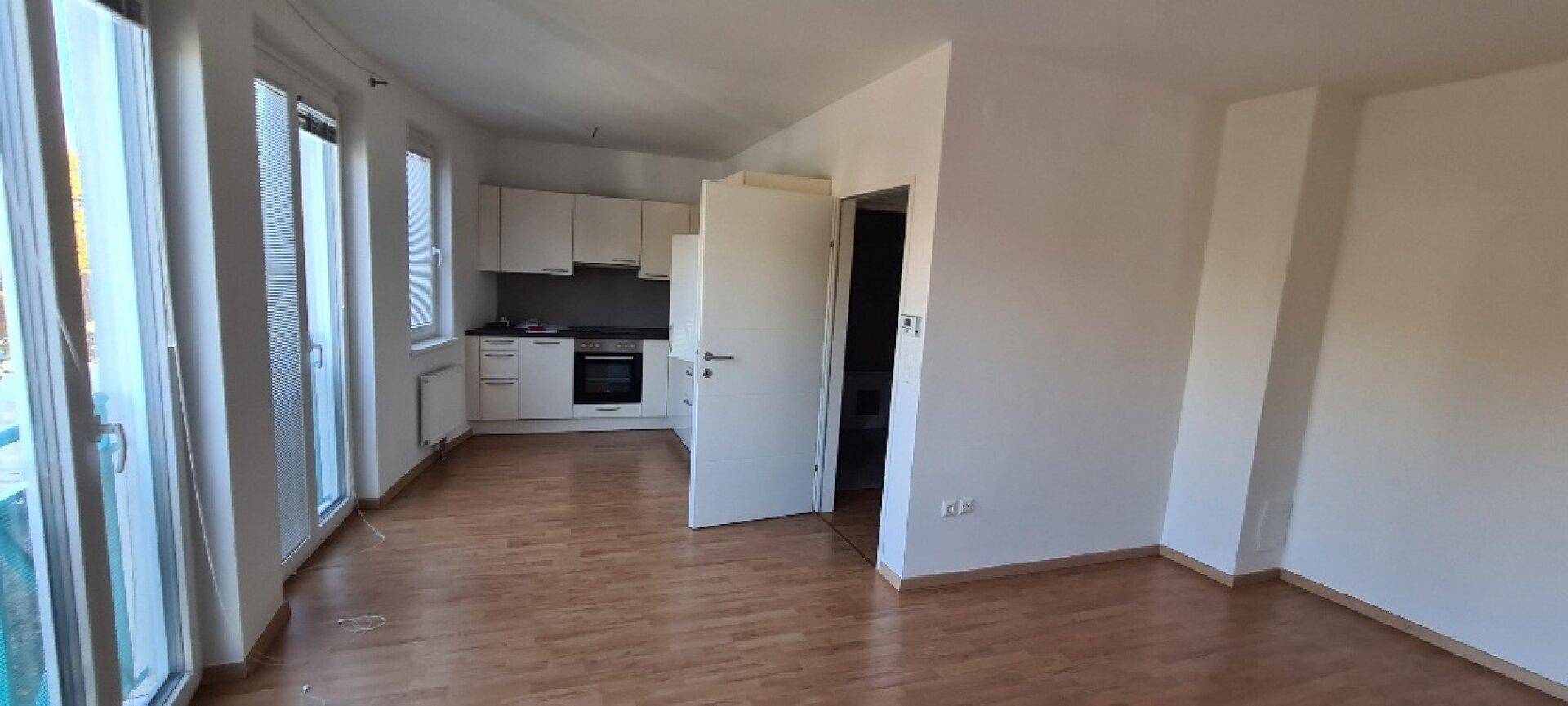 Offener Wohnbereich mit Holzboden, großen Fenstern und Blick auf die angrenzende Küche.