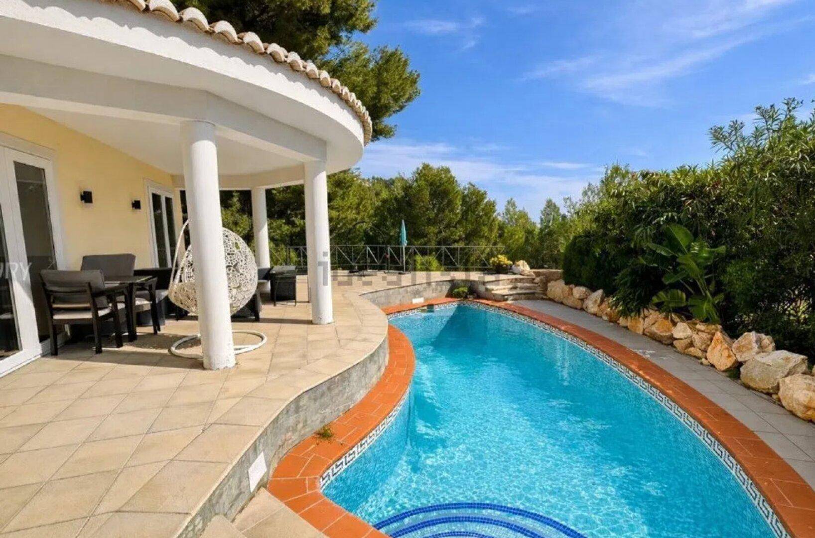 Geschwungener Poolbereich mit gefliester Terrasse und überdachtem Sitzbereich am Haus.