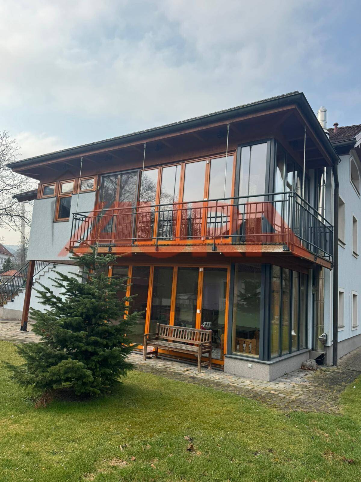 Ansicht des Hauses mit verglastem Anbau, Balkon und gepflegtem Gartenbereich mit Nadelbaum.