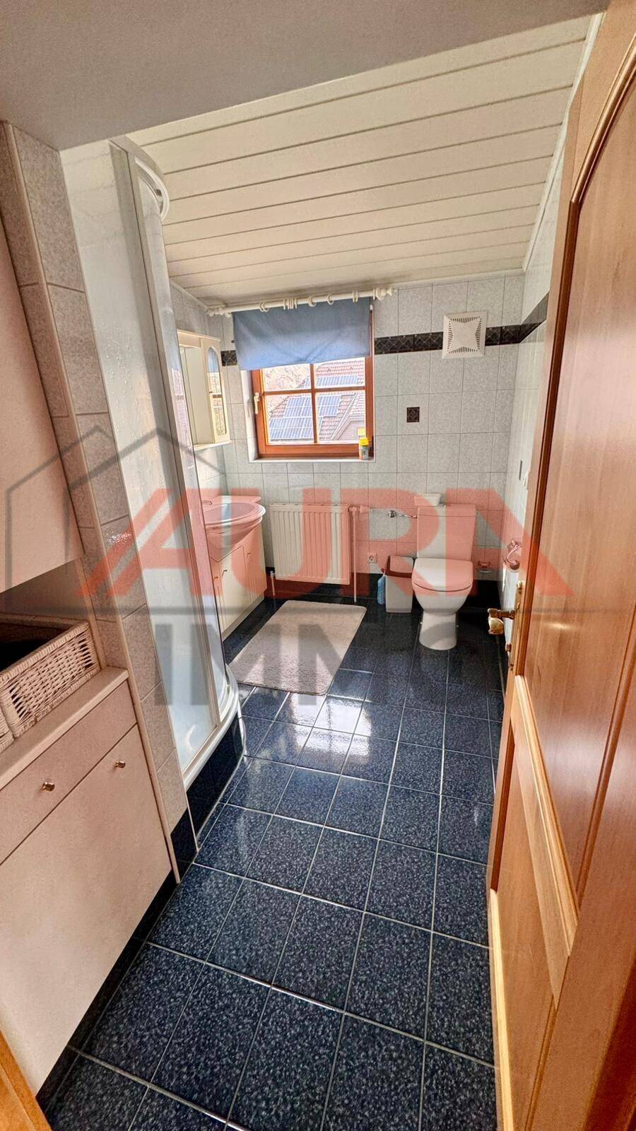Badezimmer mit Duschkabine, WC, Waschbeckenunterschrank und Fliesenboden, mit Fenster.