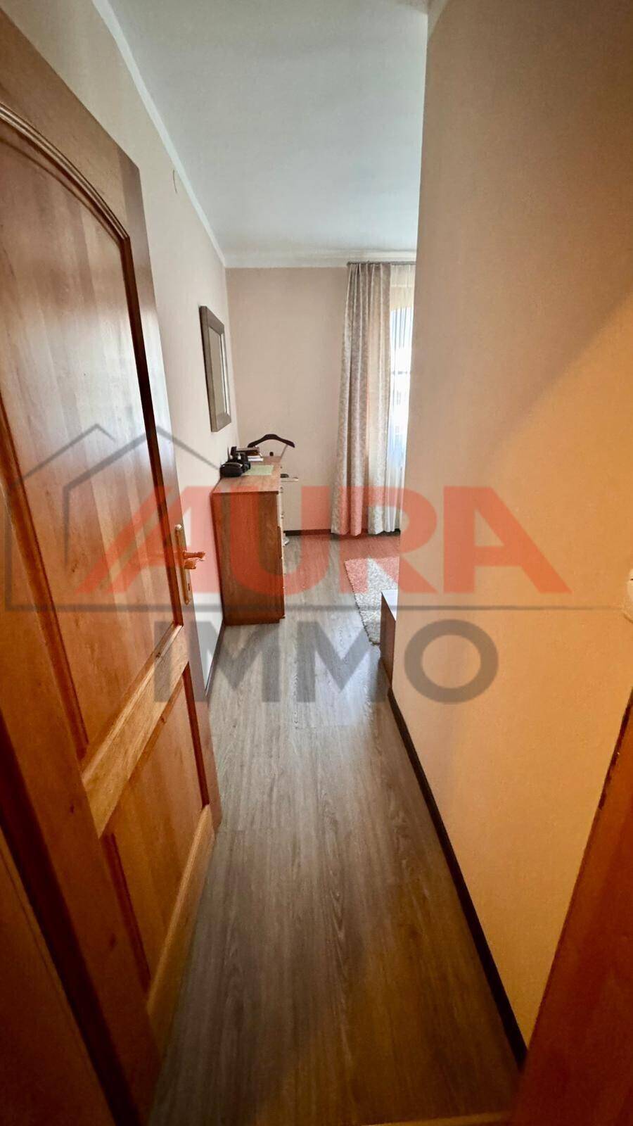 Schmaler Flur mit Laminatboden, der zu einem hellen Zimmer mit Vorhängen führt.