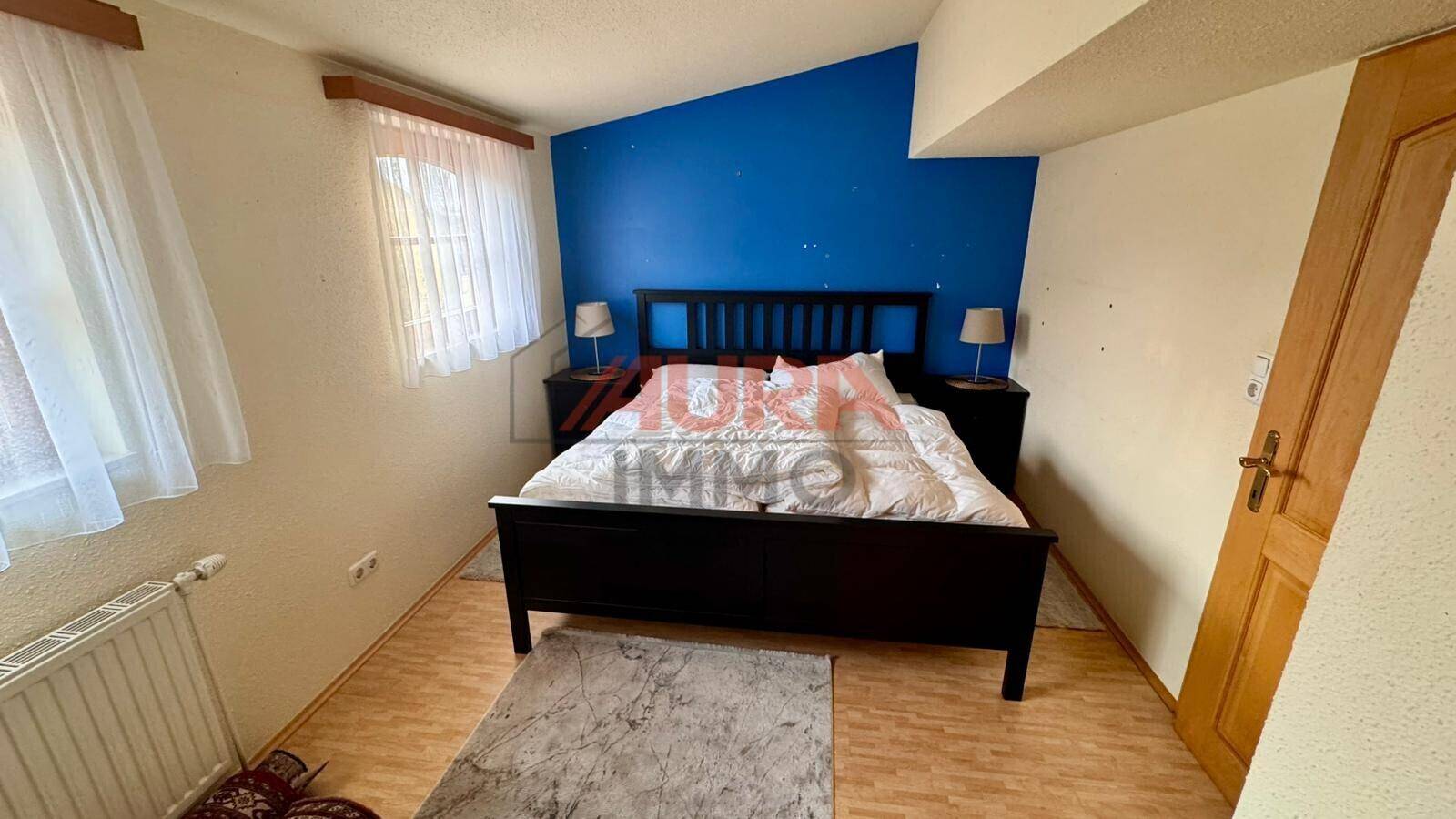 Schlafzimmer mit Doppelbett, blauer Akzentwand und Dachschräge, hell durch ein Fenster.