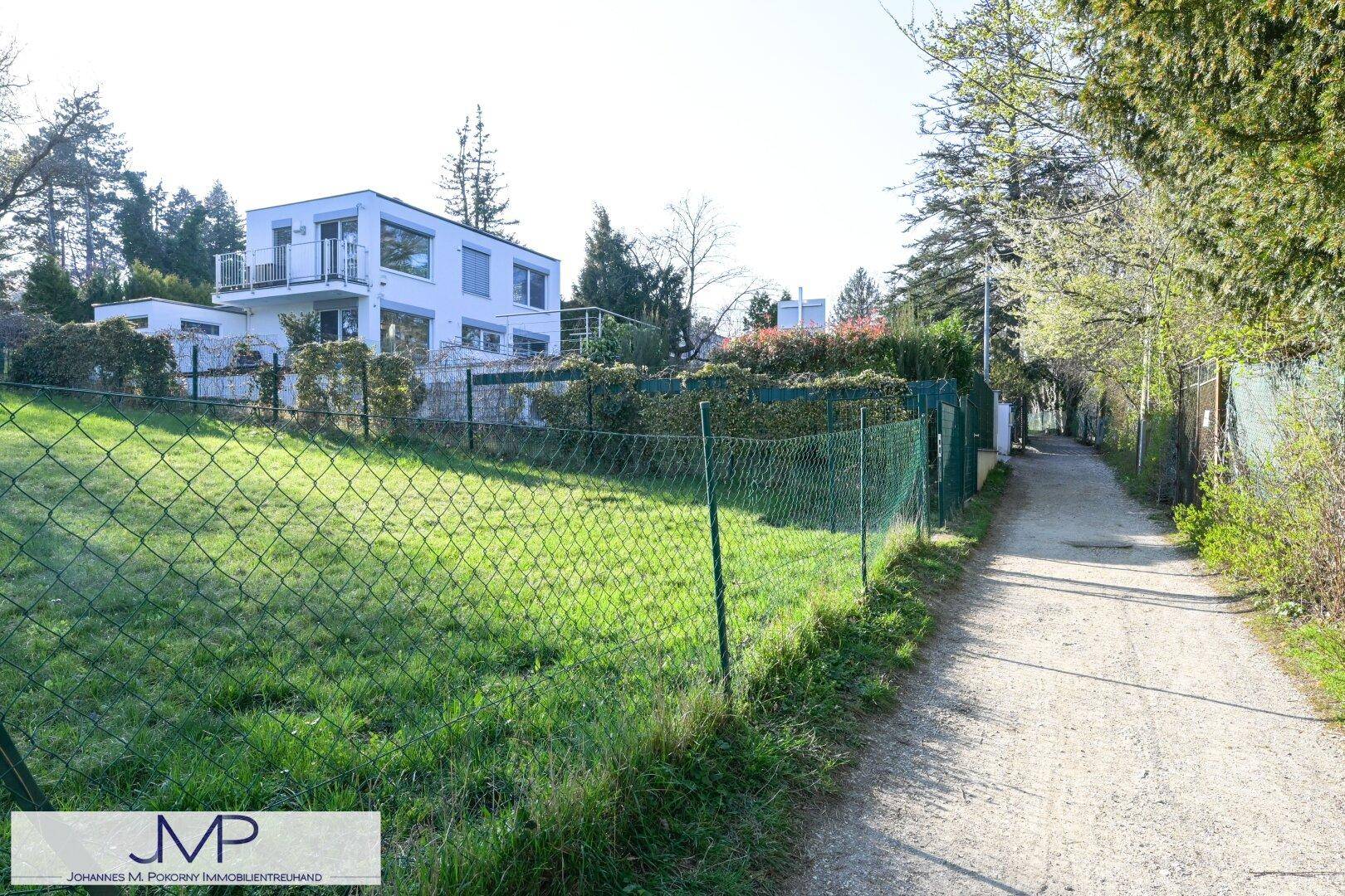 Ein Feldweg neben einem Zaun und einer Rasenfläche mit einem modernen Haus im Hintergrund.