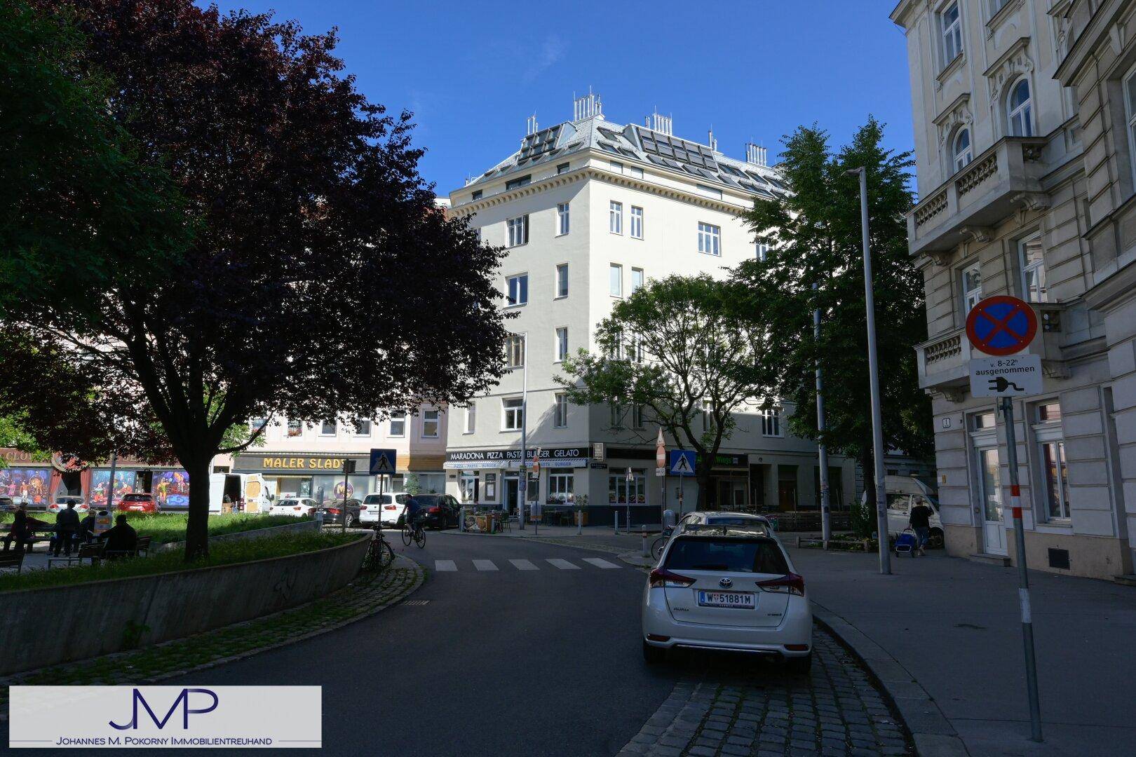 Straßenansicht eines Eckgebäudes in einer belebten Gegend mit Bäumen und parkenden Fahrzeugen.