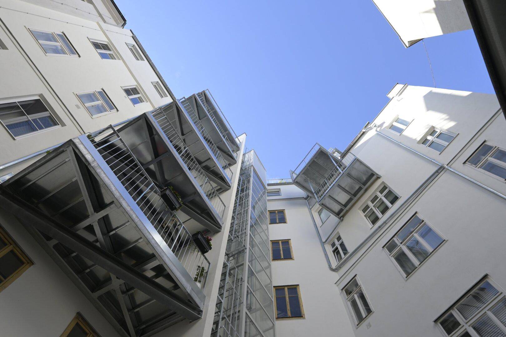 Blick in einen Innenhof mit modernen Balkonen, einer Glasfassade und einem sichtbaren Aufzugsschacht.