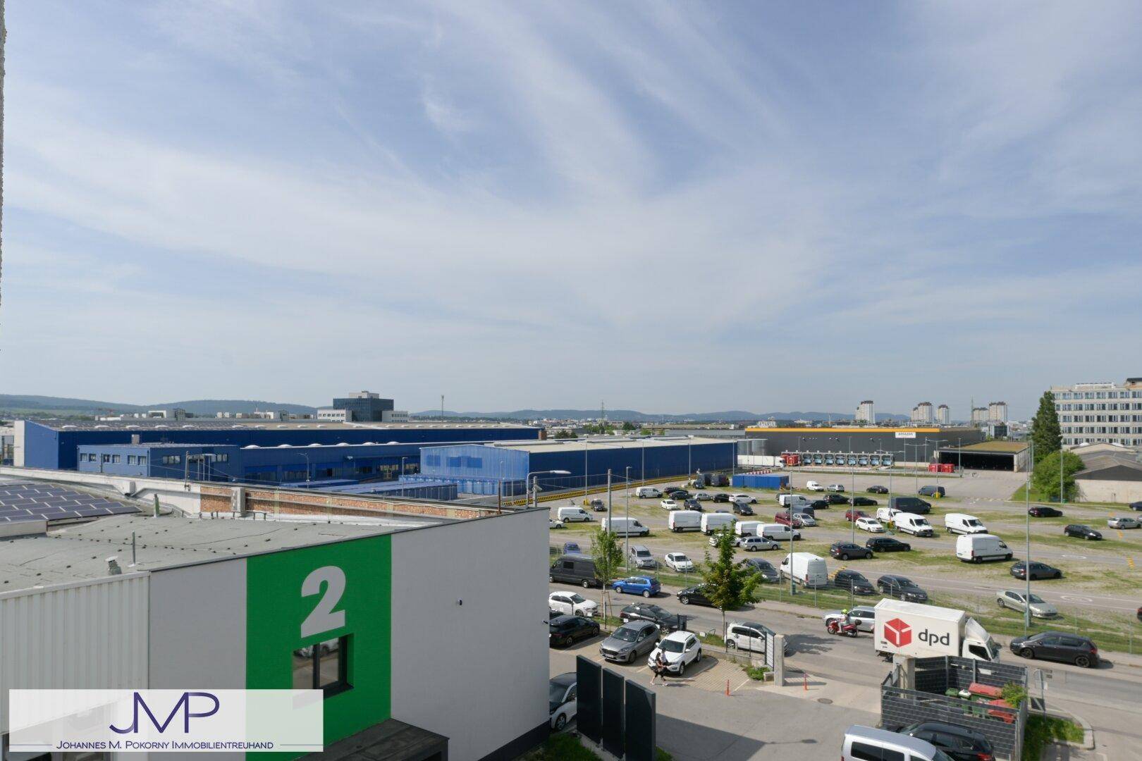 Blick über ein Industriegebiet mit großen Lagerhallen, vielen Fahrzeugen und Bergen im Hintergrund.