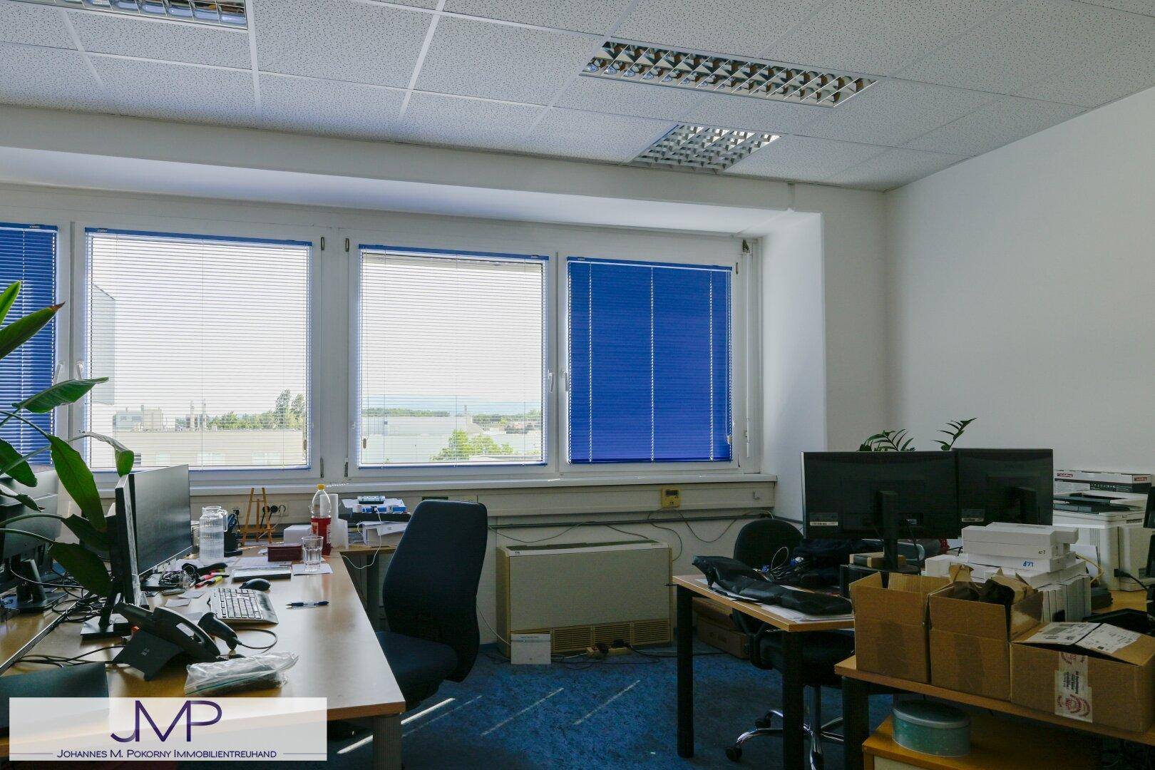 Heller Büroraum mit mehreren Arbeitsplätzen, Computern und blauen Jalousien an den Fenstern.