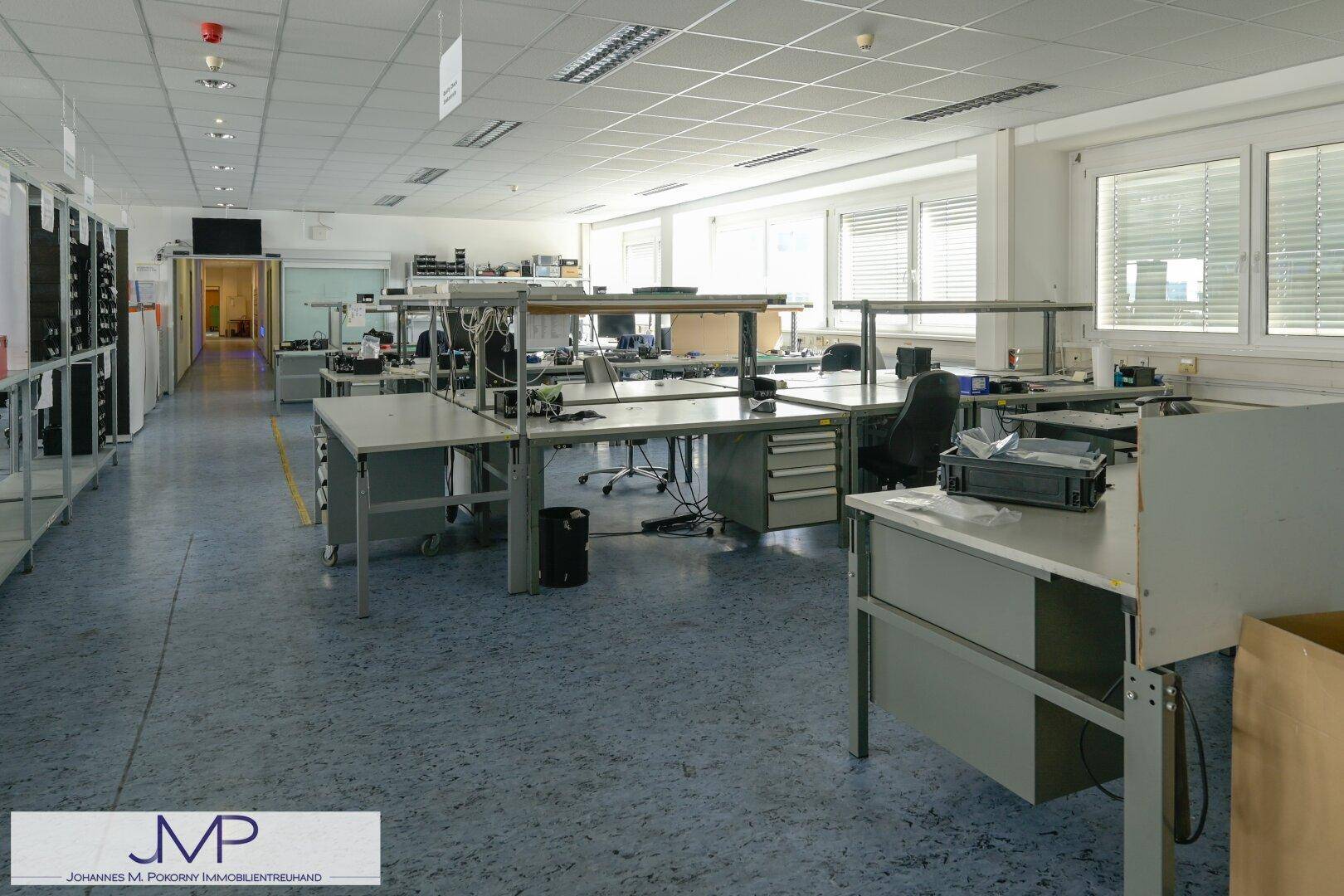 Großer, offener Bürobereich oder Werkstatt mit vielen Arbeitstischen und Fenstern für Tageslicht.