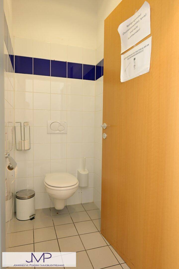 Toilettenkabine mit Wand-WC, Fliesen an Wänden und Boden, sowie einem kleinen Mülleimer.