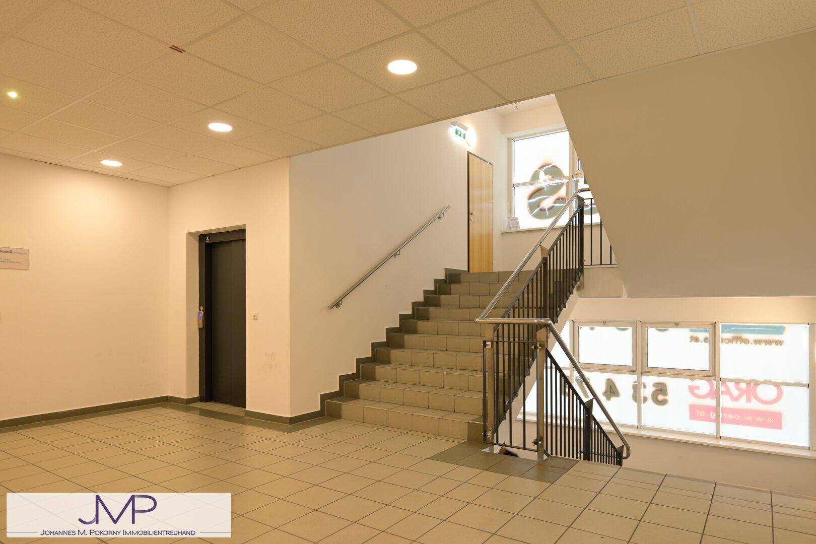 Treppenhaus mit Fliesenboden, Metallgeländer und einem Aufzug im Hintergrund.