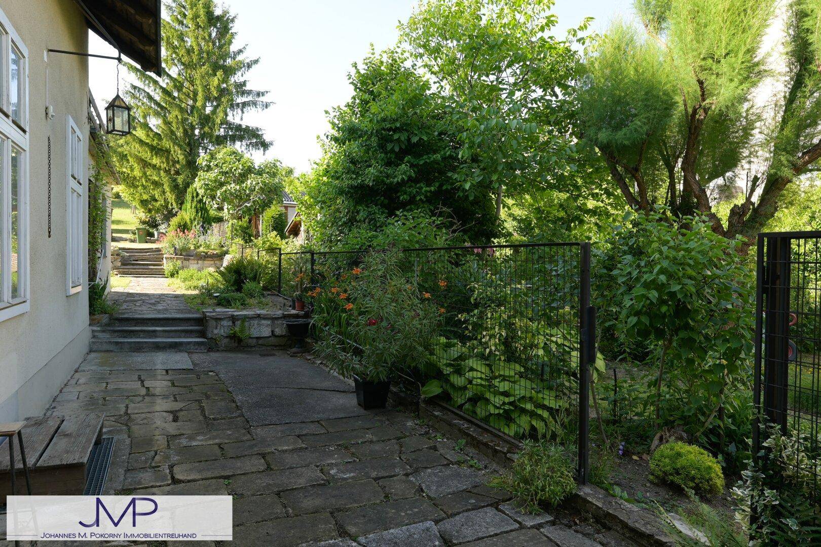 Gepflasterte Terrasse neben dem Haus, umgeben von üppigem Grün und einem Zaun.