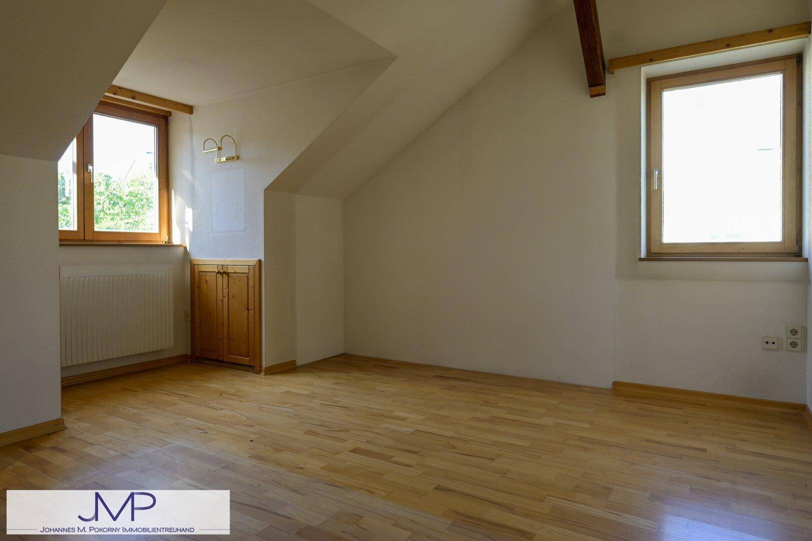 Helles Schlafzimmer im Dachgeschoss mit Holzboden, schrägen Wänden und zwei Fenstern.