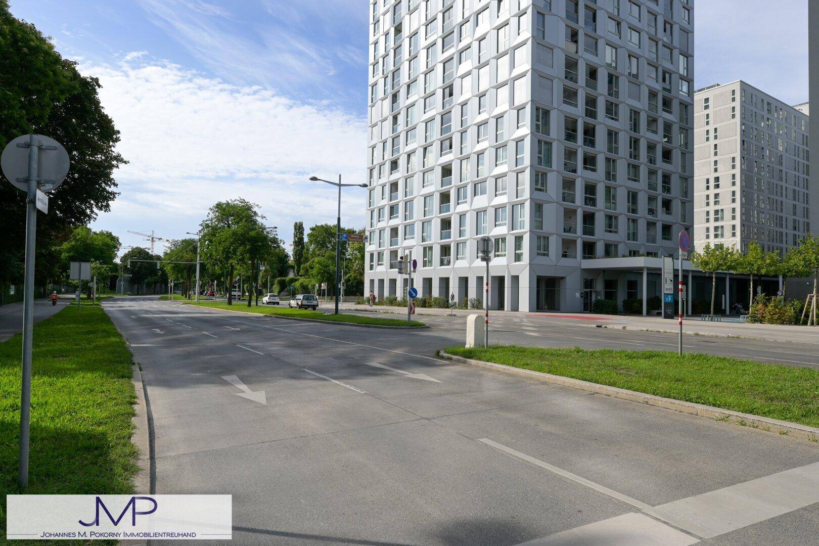 Breite Straße mit Grünstreifen und dem modernen Hochhaus im Hintergrund unter blauem Himmel.