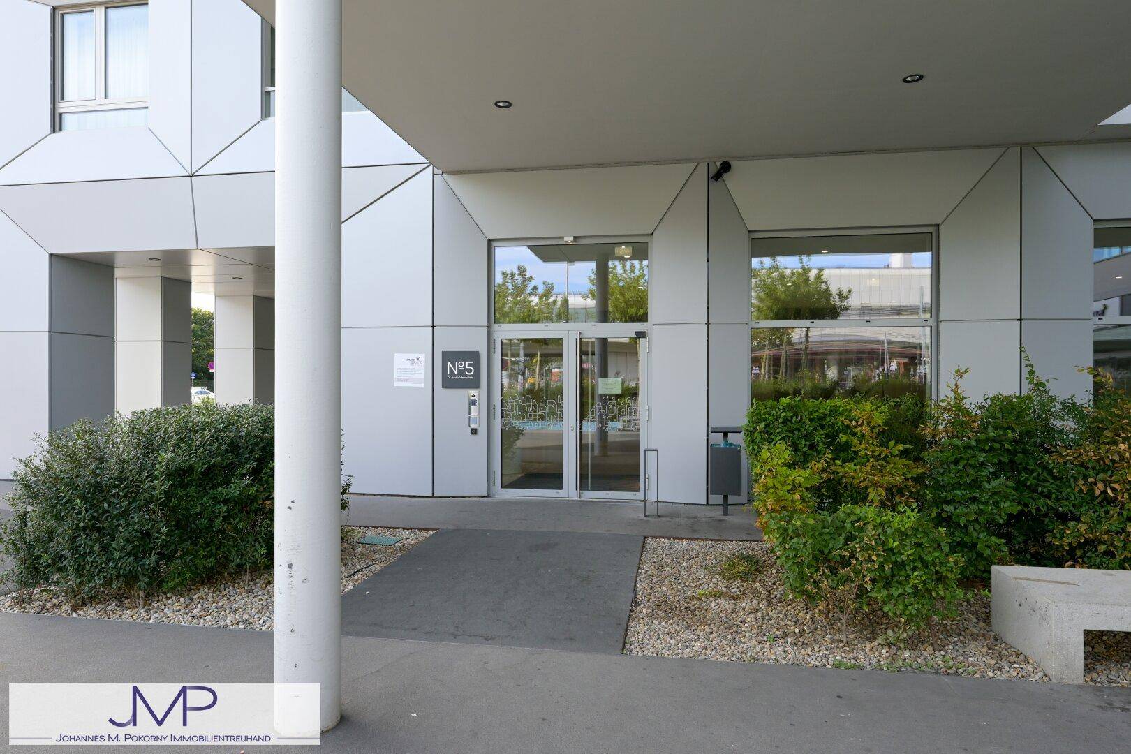 Hauptzugang eines modernen Gebäudes mit Glastüren und gepflegten Sträuchern im Eingangsbereich.