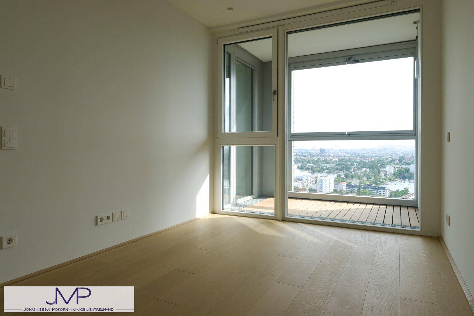 Leeres Zimmer mit hellem Holzboden und Zugang zu einer Loggia mit Stadtblick.