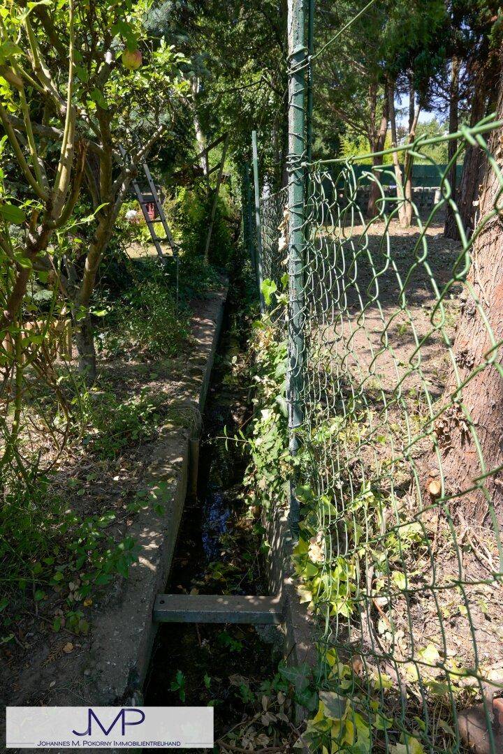 Ein schmaler Wassergraben mit Betonbegrenzung und einem Zaun in einem grünen Gartenbereich.