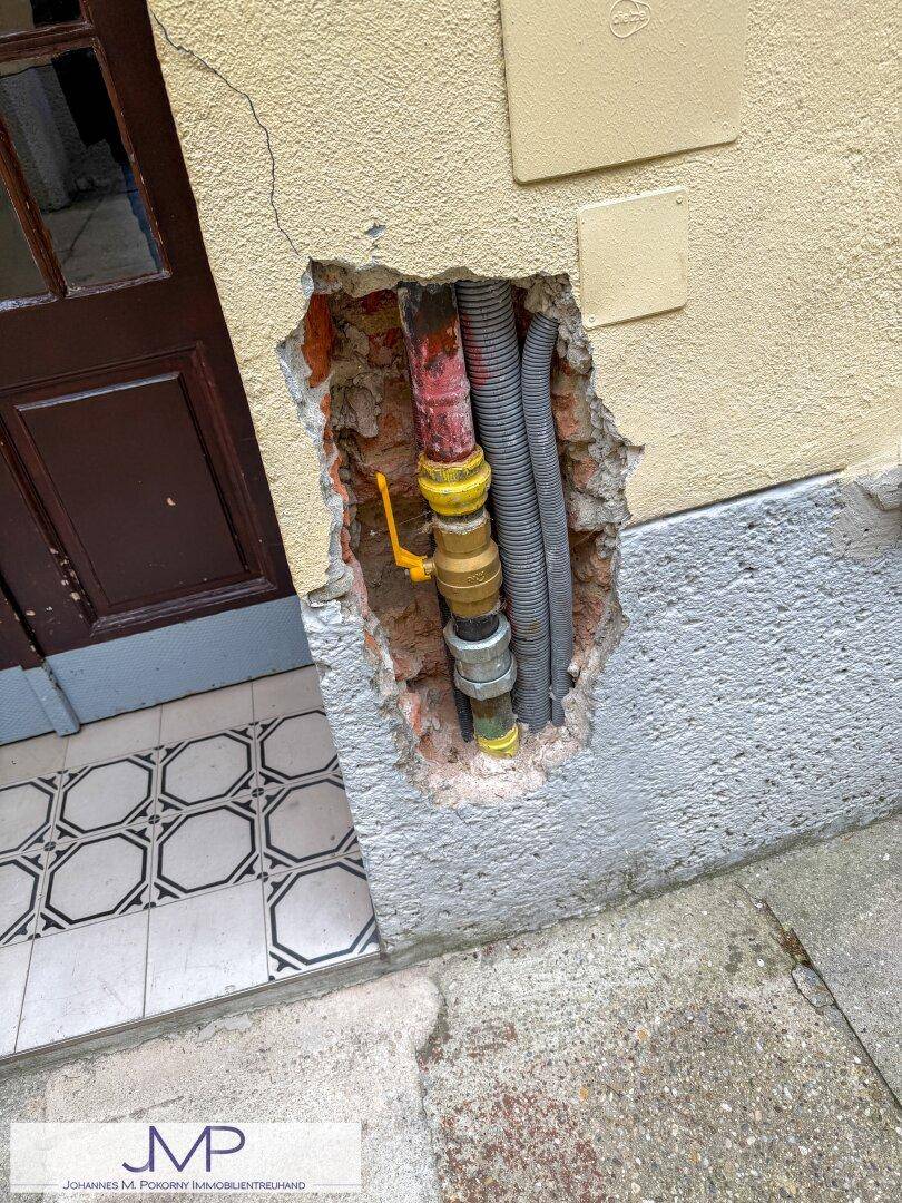 Detailaufnahme einer Wand mit freigelegten Rohren und Leitungen neben einem Hauseingang.