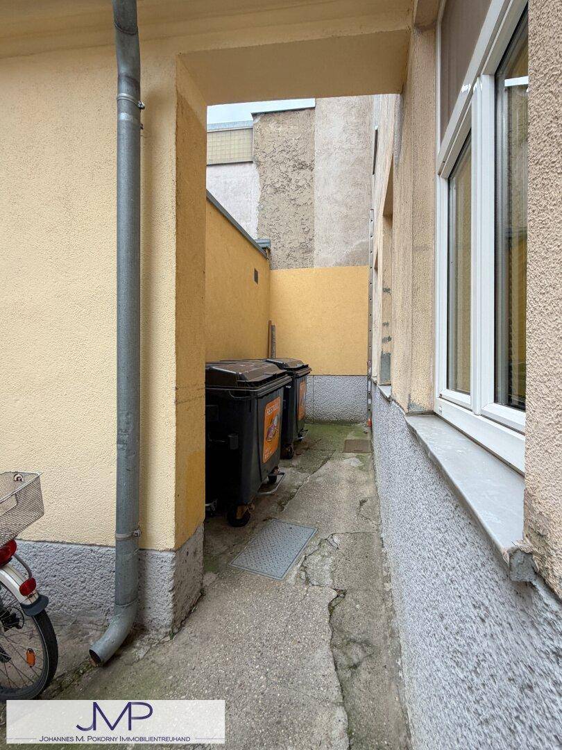 Schmaler Durchgang zwischen Gebäuden mit Mülltonnen und einem Fallrohr an der Wand.