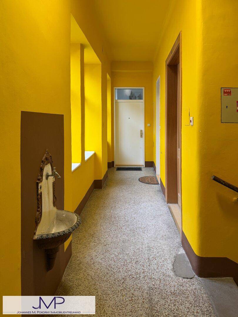 Gelb gestrichener Flur mit Terrazzoboden, einem historischen Wandbrunnen und einer Eingangstür.