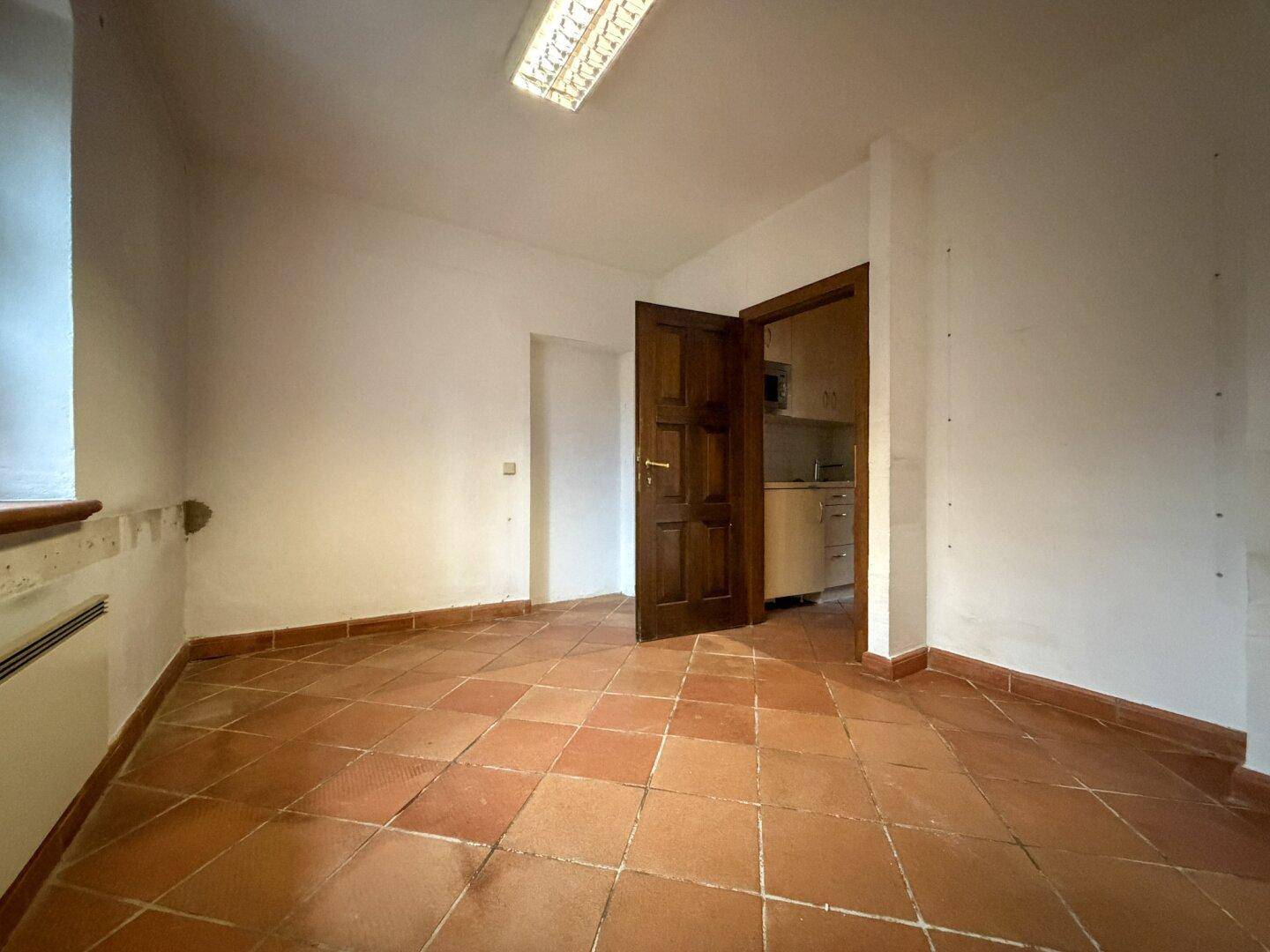 Leerer Raum mit Terrakottafliesen und einem Blick durch die offene Tür in eine kleine Küche.