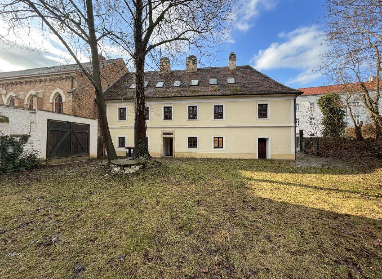 Rückansicht des historischen Gebäudes vom Garten aus mit Bäumen und einem Nebengebäude mit Holztoren.