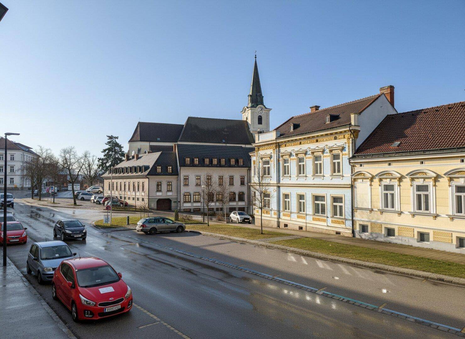 Blick auf eine belebte Straße mit geparkten Autos, historischen Gebäuden und einem Kirchturm im Hintergrund.