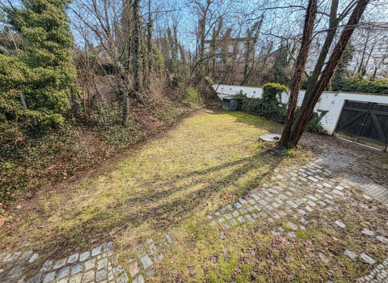 Privater Gartenbereich mit Rasenfläche und gepflasterten Wegen, umgeben von Bäumen und einer Mauer.