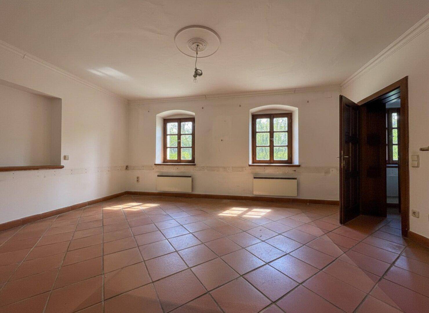 Geräumiger, leerer Raum mit Terrakottafliesen, zwei Fenstern und einem Deckenstuckelement.
