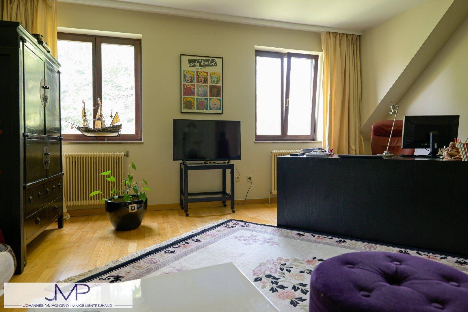 Helles Arbeitszimmer mit einem großen Schreibtisch, Holzboden und Blick ins Grüne durch die Fenster.